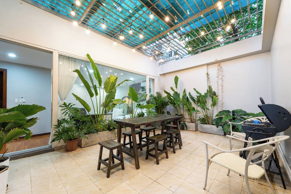 Eine überdachte Terrasse mit einem Holztisch und -stühlen, umgeben von Blumentöpfen und einem Grill.