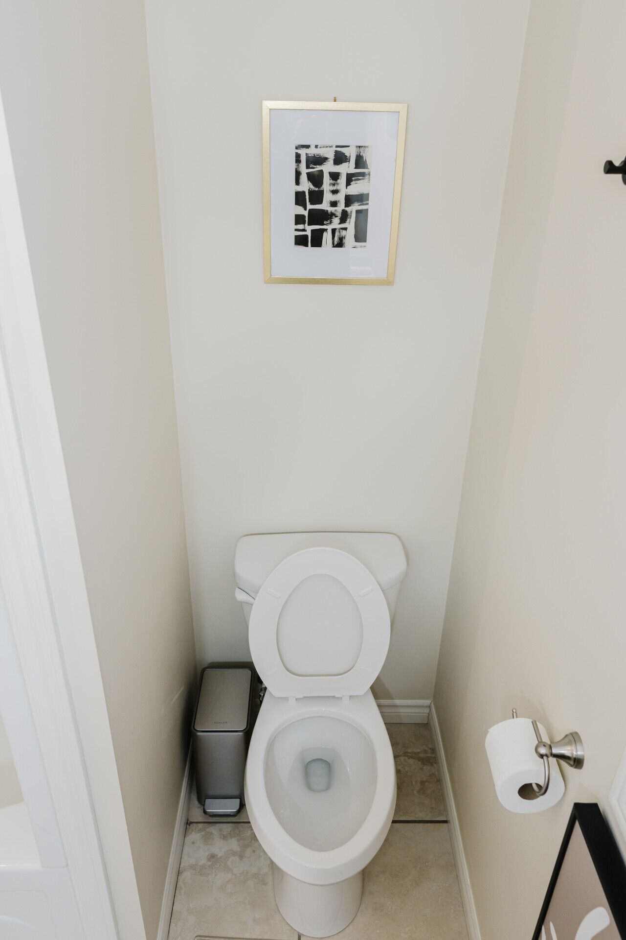 A small bathroom with a toilet, a trash bin, a toilet paper holder, and a framed picture on the wall.