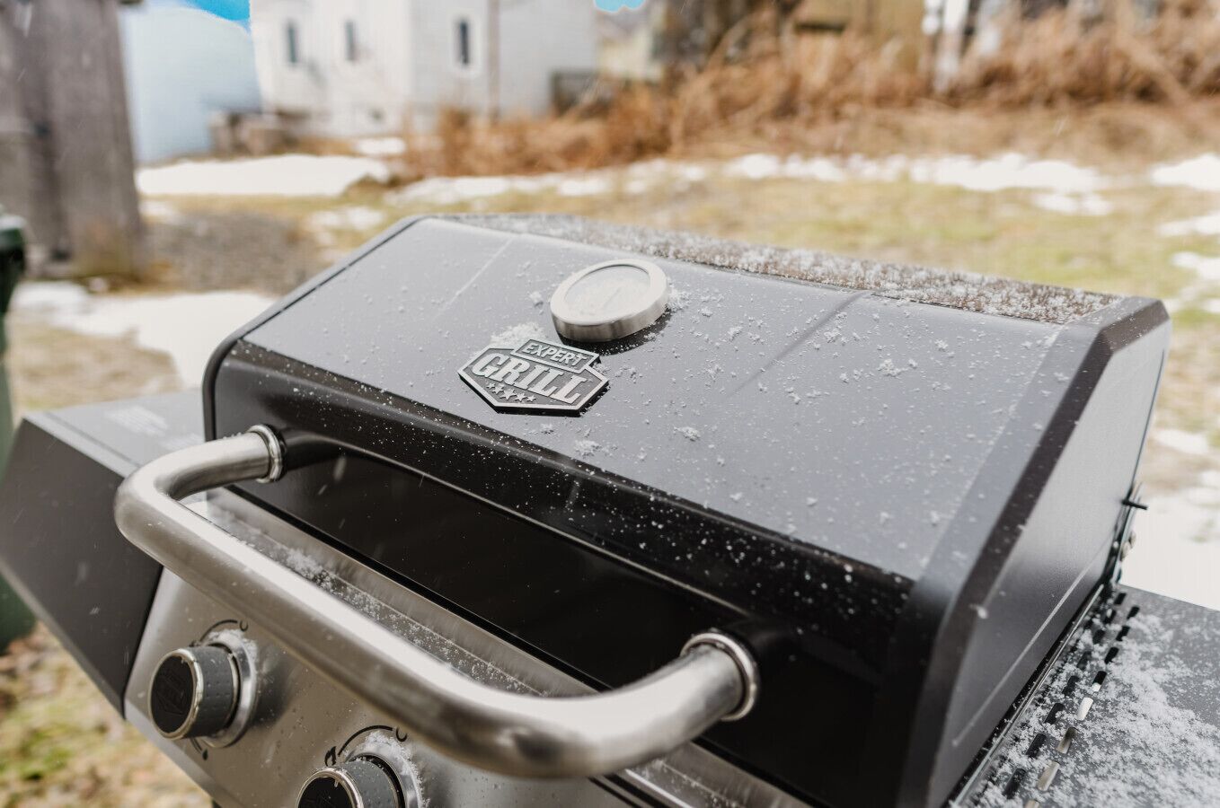 Ein Grill mit Griff und Reglern, mit dem Schriftzug 'Expert Grill', bedeckt mit Schnee.