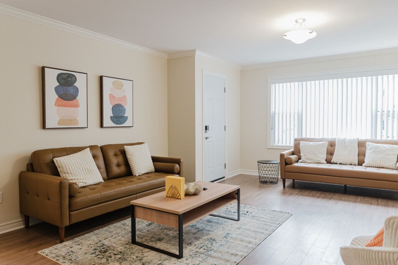 Un salon avec un canapé en cuir marron, une table basse en bois et un canapé beige avec des coussins blancs. Deux tableaux abstraits ornent le mur, ainsi qu’une porte blanche et une fenêtre avec des stores.