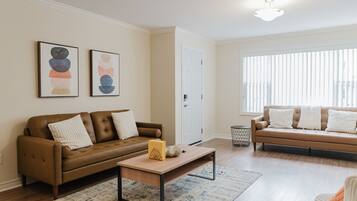 Un salon avec un canapé en cuir marron, une table basse en bois et un canapé beige avec des coussins blancs. Deux tableaux abstraits ornent le mur, ainsi qu’une porte blanche et une fenêtre avec des stores.