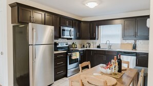 A modern kitchen with dark cabinets, stainless steel appliances, a dining table with chairs, and a window with blinds. - Garden Suite | Modern 2BR with Private Yard (Sydney)