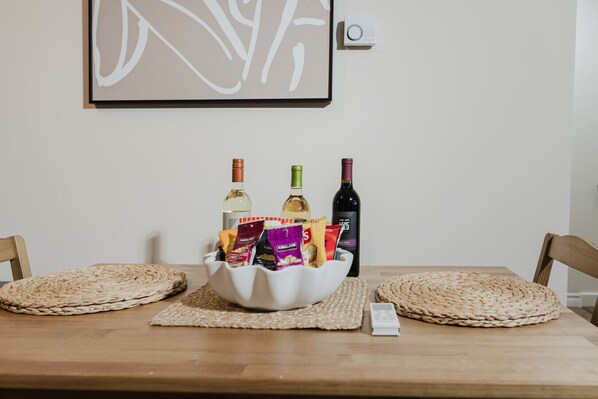 Une table à manger avec un bol blanc contenant des amuse-gueules et deux bouteilles de vin, un tableau blanc orné d’une œuvre d’art abstraite et un thermostat fixé au mur.