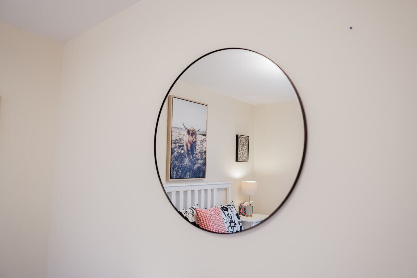 Una habitación con un espejo ovalado grande en la pared, un radiador debajo, un cuadro enmarcado y una lámpara sobre una mesita.