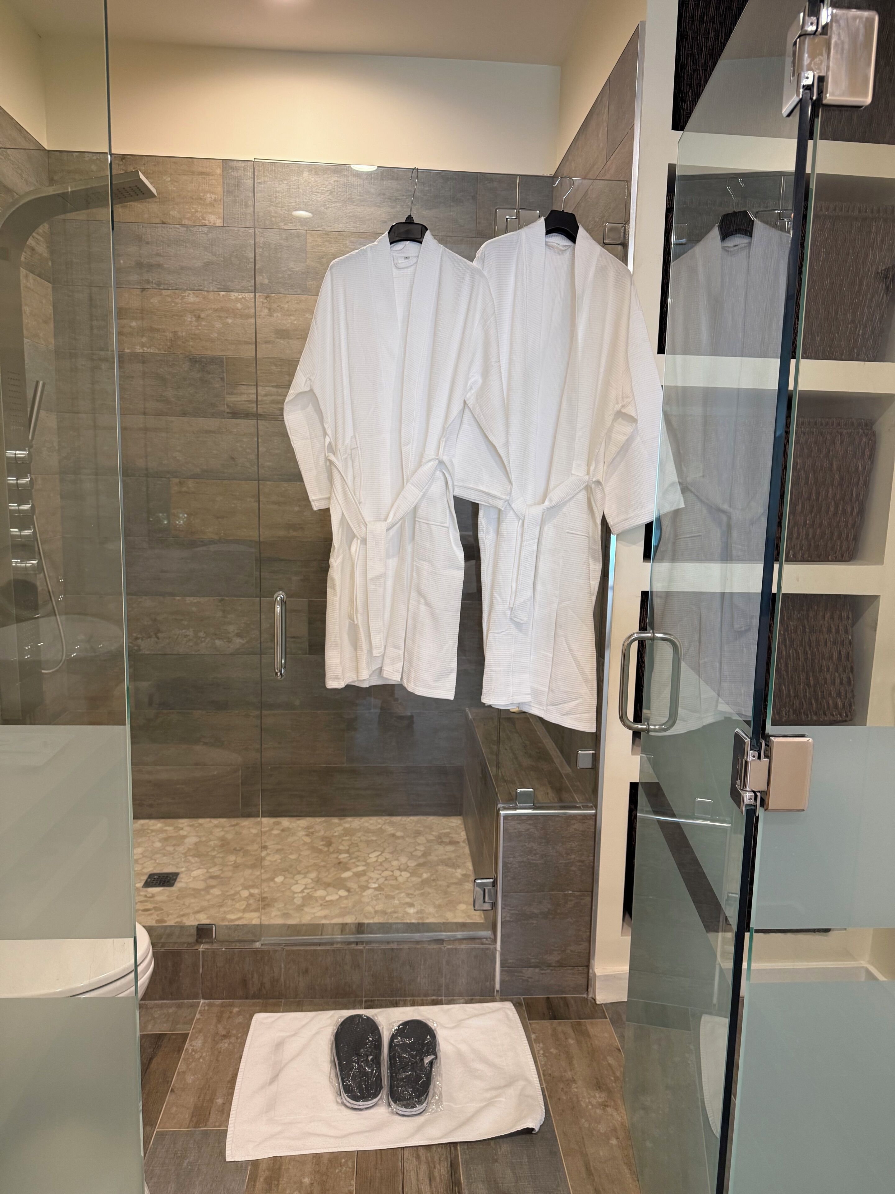 Een moderne badkamer met een glazen douchecabine, twee witte badjassen aan een rek en een paar slippers op een handdoek op de vloer.