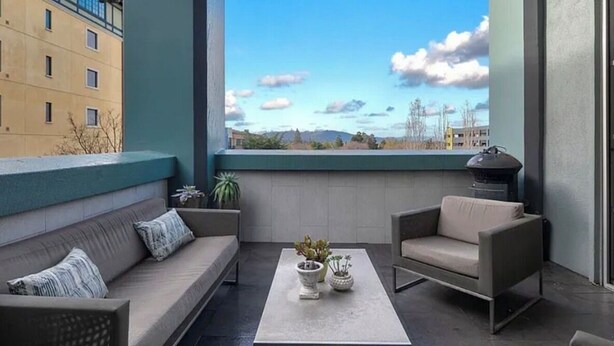Un balcón con sofá, sillón y mesa, y vista a edificios y un cielo despejado.