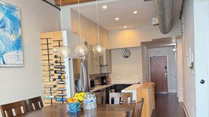 Un comedor con una mesa y sillas de madera, una cocina al fondo y conductos visibles en el techo.