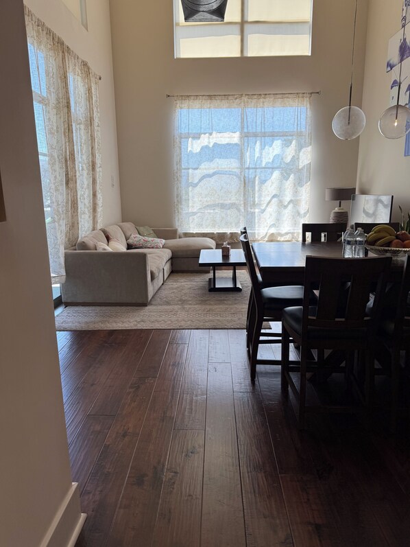 A living room with a sofa, a dining table with chairs, and a large window with curtains.