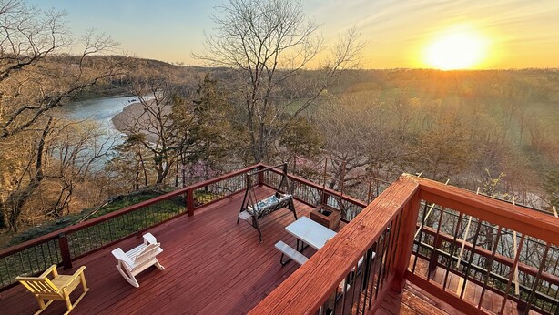 Een houten terras met buitenmeubilair met uitzicht op een rivier en een bebost landschap tijdens de zonsondergang.