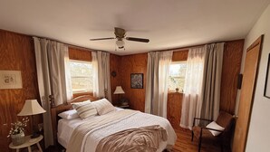 Een slaapkamer met een bed, een stoel, een nachtkastje, een lamp, een plafondventilator en een raam met gordijnen.