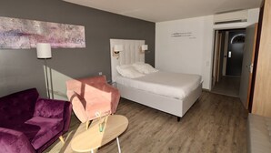 A hotel room with a bed, a pink armchair, a small round table, a floor lamp, and a painting on the wall. - Le Jardin d'Eden JE112 - Suite Junior PMR Luxe - 2 people (AGDE)