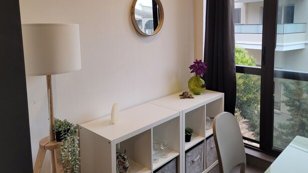 A modern room with a white console table, a mirror, a vase with flowers, a lamp, and a view of buildings through a window. - Spacious studio in residence with swimming pool close to all amenities (Mougins)