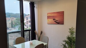 A room with a dining table, chairs, a window with curtains, a painting on the wall, and a potted plant. - Spacious studio in residence with swimming pool close to all amenities (Mougins)