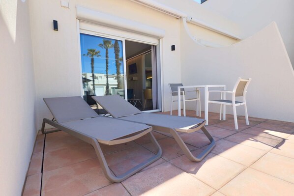 A sunlit outdoor area with two lounge chairs and a small table, a sliding glass door leading to an interior space, and a tiled floor.