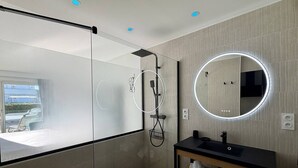 A modern bathroom with a glass shower enclosure, a round mirror with LED lights, a black sink, and a wooden vanity. - Le Jardin d'Eden 104 - Suite Junior Luxe - 2 people (AGDE)