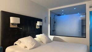 A modern hotel room with a large bed, two wall-mounted lamps, a large mirror, and a shower area. - Le Jardin d'Eden 104 - Suite Junior Luxe - 2 people (AGDE)