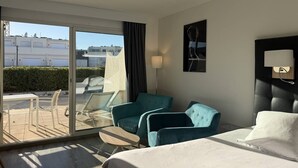 A hotel room with a bed, two armchairs, a table, a lamp, and a large window with a view of buildings. - Le Jardin d'Eden 104 - Suite Junior Luxe - 2 people (AGDE)