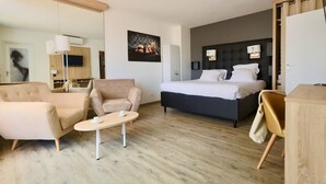 A modern hotel room with a bed, a sofa, a chair, a small table, and a desk. - Le Jardin d'Eden 211 - Suite Junior Premium - 2 people (AGDE)
