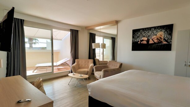 A hotel room with a bed, two armchairs, a small table, a TV, and a balcony with a sun lounger. - Le Jardin d'Eden 211 - Suite Junior Premium - 2 people (AGDE)