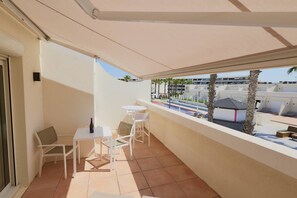 A balcony with white furniture, a bottle of wine, and a view of a pool and buildings.