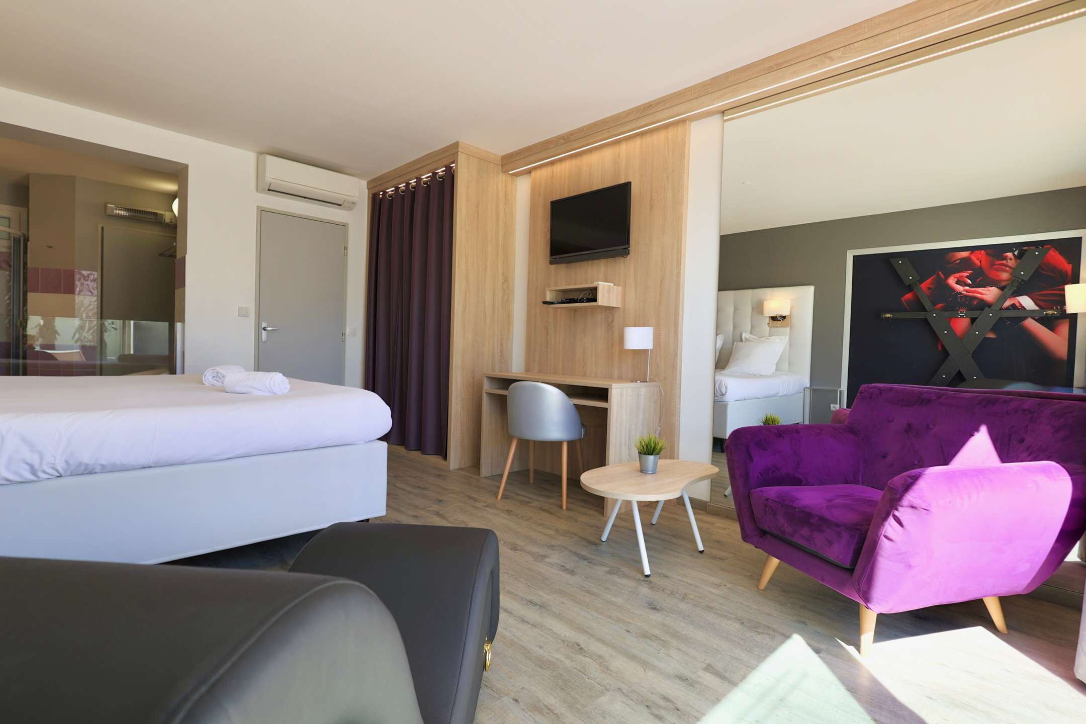 A hotel room with a bed, a desk, a purple armchair, and a television.