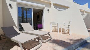 Un área de estar exterior moderna con dos sillones de descanso y una mesa, una puerta corrediza de vidrio que da a un espacio interior y una pared exterior blanca.