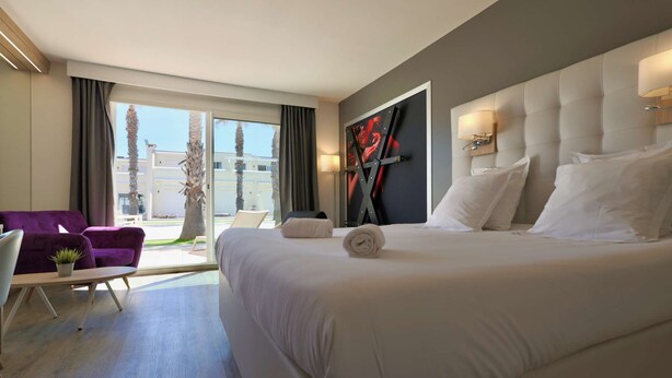 Habitación de hotel con una cama grande, un sillón morado, una mesita con una planta y vistas al exterior.
