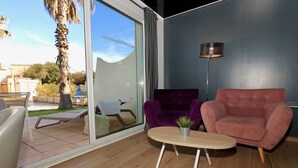 Una sala de estar moderna con un ventanal que da a un espacio exterior, un sofá morado, un sofá rosa, una mesita con una planta y una lámpara de pie.