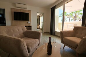 Habitación de hotel con sofá, televisión montada en la pared, una botella de vino sobre una mesa y vistas al exterior.