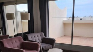 Habitación de hotel con cama, dos sillones, una mesita y ventanas amplias con vistas al cielo y edificios.
