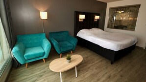 Une chambre d’hôtel avec un lit, deux fauteuils turquoise, une petite table ronde avec une plante et des lampes fixées au mur.