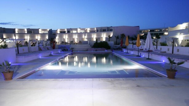 Une piscine entourée de chaises longues et de parasols, elle-même bordée d’un bâtiment blanc doté de nombreux balcons.