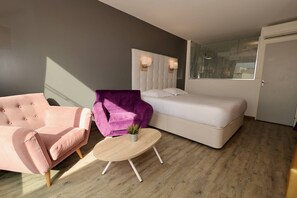 Une chambre à coucher moderne avec un lit, un fauteuil rose, un fauteuil violet et une petite table ronde agrémentée d’une plante.