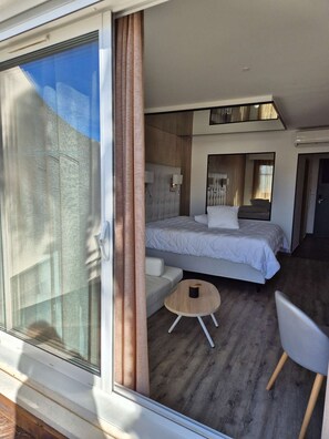 A modern hotel room with a large sliding glass door, a bed with a headboard, a small round table, and a chair. - Le Jardin d'Eden - Suite Junior Luxe 139 with large terrace - 2 people (AGDE)