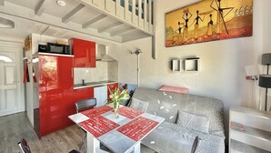 A modern kitchen with red cabinets, a dining area with a table set for two, and a living area with a sofa and a painting on the wall. - CAP D'AGDE - MONT ST MARTIN // Réf 0314 (CAP D'AGDE)