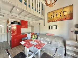 A modern kitchen with red cabinets, a dining area with a table set for two, and a living area with a sofa and a painting on the wall. - CAP D'AGDE - MONT ST MARTIN // Réf 0314 (CAP D'AGDE)
