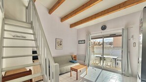 A room with a staircase, a sofa, a table, a sliding glass door leading to an outdoor area with chairs, and a clock on the wall. - CAP D'AGDE - Richelieu // Réf 0309 (CAP D'AGDE)