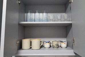 A cabinet with three shelves; the top shelf holds clear glasses, the middle shelf has a row of clear glasses, and the bottom shelf contains decorative mugs. - Business-Ready Studio in New Kingston Fast WiFi + AC (Kingston)