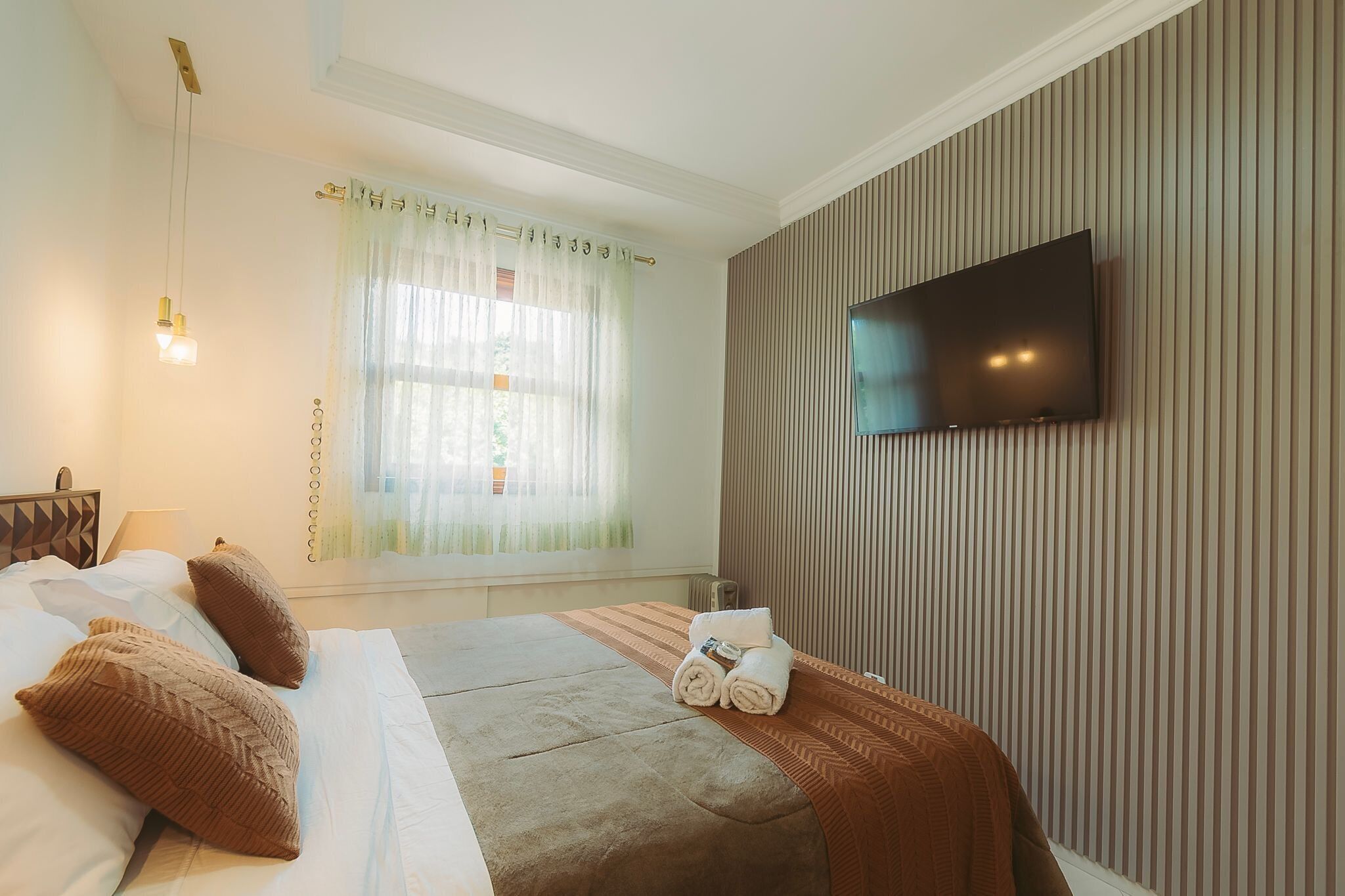 A hotel room with a bed, a TV mounted on the wall, a window with curtains, and a bedside table with a lamp.