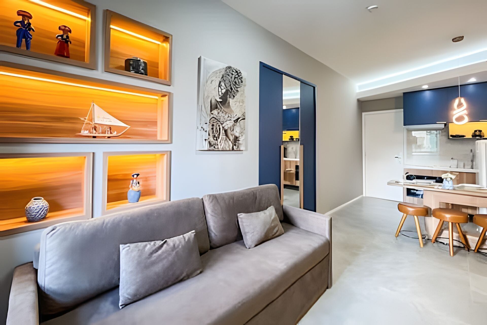 Una sala de estar moderna con un sofá gris, estanterías de madera con objetos decorativos, y una zona de cocina con una mesa de comedor y taburetes.