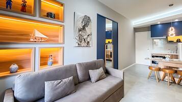 Una sala de estar moderna con un sofá gris, estanterías de madera con objetos decorativos, y una zona de cocina con una mesa de comedor y taburetes.