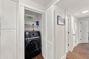 A laundry room with a front-loading washer and dryer, shelves with laundry supplies, and a door leading to another room. - BG Beach Club 3E | 3BR/2BA Boca Grande Condo (Boca Grande)