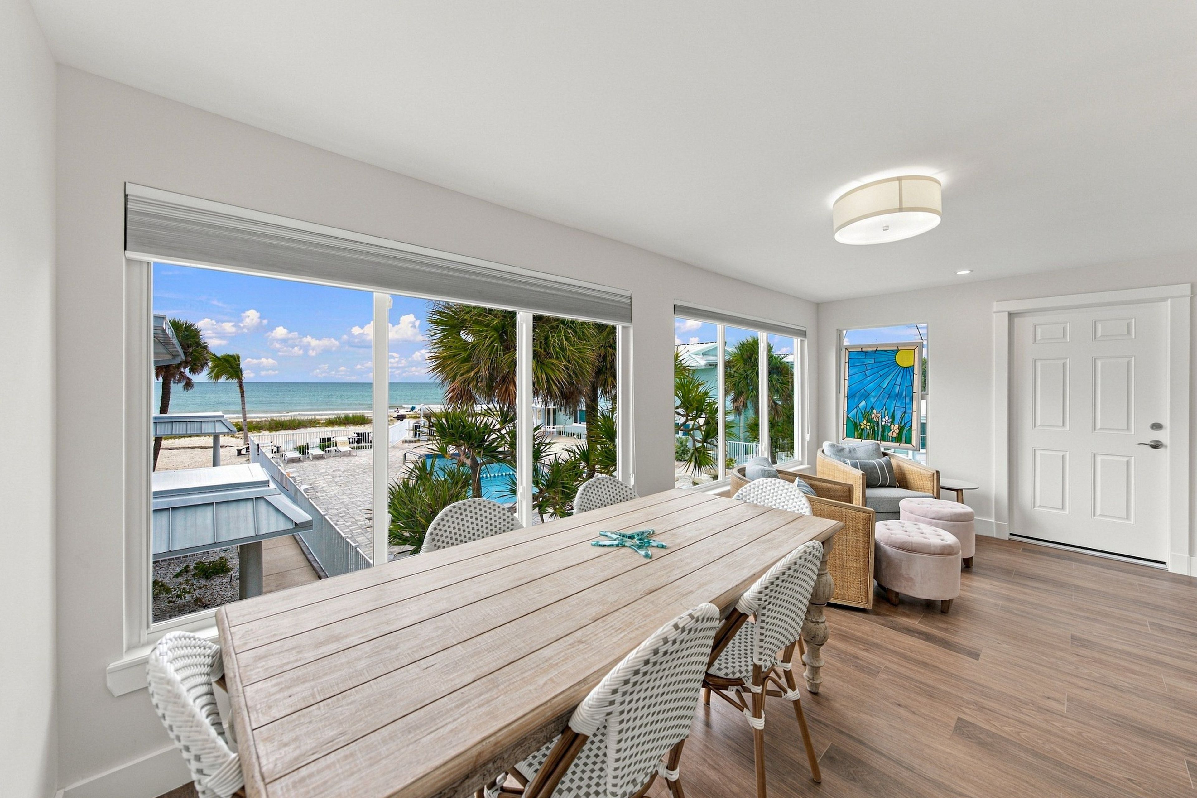 A spacious room with a large dining table, wicker chairs, and a view of a beach and pool.