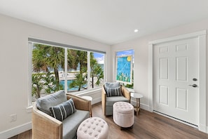 A room with a view of a pool and palm trees, furnished with a sofa, ottoman, and chair. - BG Beach Club 3E | 3BR/2BA Boca Grande Condo (Boca Grande)