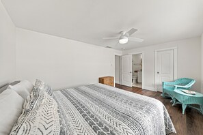 A bedroom with a bed, a ceiling fan, a wicker chair, and a door leading to a bathroom. - BG Beach Club 3E | 3BR/2BA Boca Grande Condo (Boca Grande)