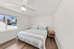A bedroom with a bed, a nightstand, a ceiling fan, and a window with a view of palm trees. - BG Beach Club 3E | 3BR/2BA Boca Grande Condo (Boca Grande)
