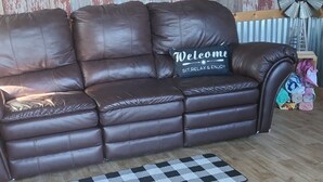 Un canapé en cuir marron agrémenté d’un coussin sur lequel est brodé le mot « Bienvenue », un tapis à carreaux et un plancher en bois.