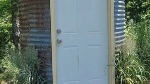 Une petite cabane rustique avec une porte blanche, une fenêtre à sommet incurvé et deux lanternes fixées au mur.