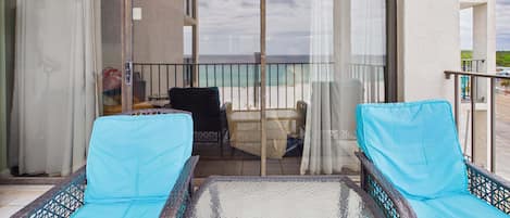 A balcony with wicker furniture, a glass table, and a view of the beach and buildings.