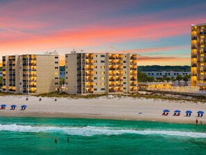 Ein Hotel direkt am Strand mit mehreren Etagen, Balkonen und einem Sandstrand mit Sonnenschirmen.